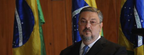 O ex-ministro Antonio Palocci sentado e uma bandeira do Brasil ao fundo