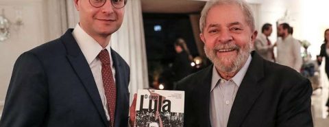 Lula usa um terno preto e camisa branca sem gravata. Lula segura um livro entregue a ele pelo advogado Cristiano Zanin, que usa um terno preto, gravata vermelha, camisa branca e óculos. Os dois estão sorrindo