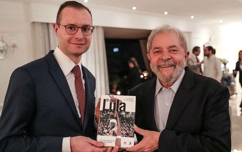 Lula usa um terno preto e camisa branca sem gravata. Lula segura um livro entregue a ele pelo advogado Cristiano Zanin, que usa um terno preto, gravata vermelha, camisa branca e óculos. Os dois estão sorrindo