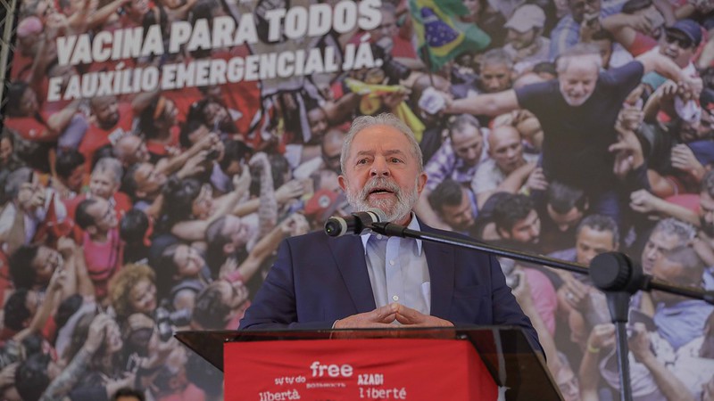 Lula veste um terno preto e camisa azul, enquanto discursa em uma tribuna com uma bandeira vermelha escrita "Lula Livre" em várias linguas. Ao fundo, um painel com uma foto de Lula cercado pelo povo