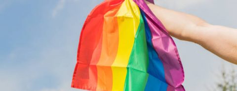 Mão segurando uma bandeira com as cores do arco íris simbolizando a comunidade LGBTQIA+