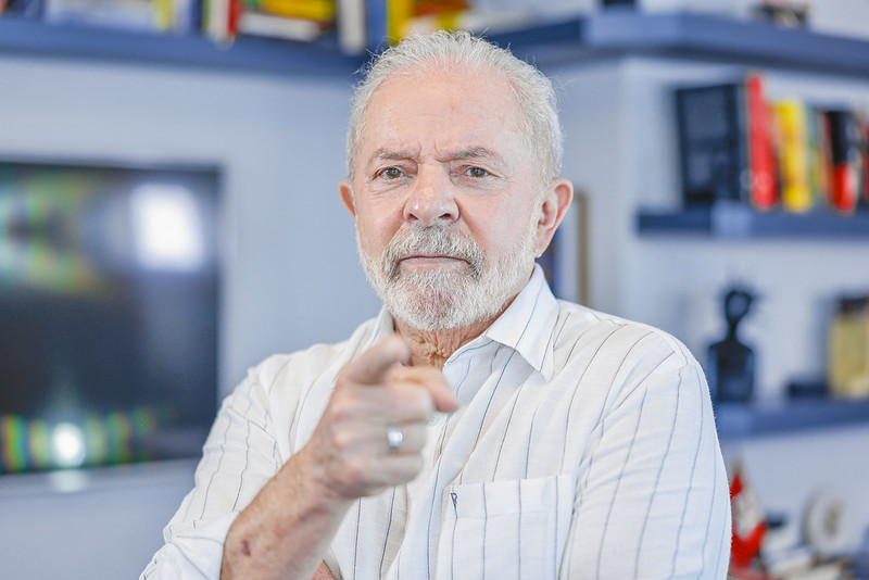 Lula, ex-presidente, veste uma camisa branca listrada, aponta o dedo indicador em direção à câmera, em retrato em plano médio