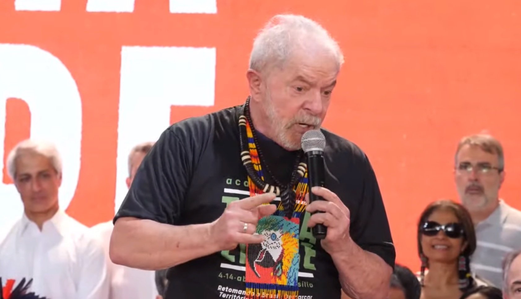 Lula participa de evento com movimentos indígenas em Brasília. Ele veste uma camiseta preta e um colar que representa a cultura indígena