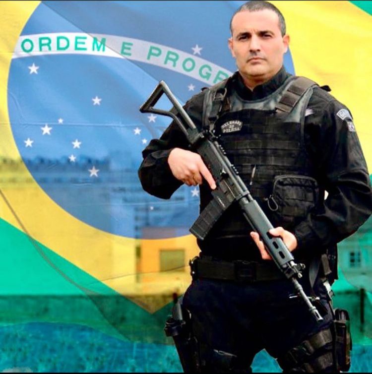 Vereador Delegado Palumbo fardado, com fuzil na mão e a bandeira do Brasil ao fundo.