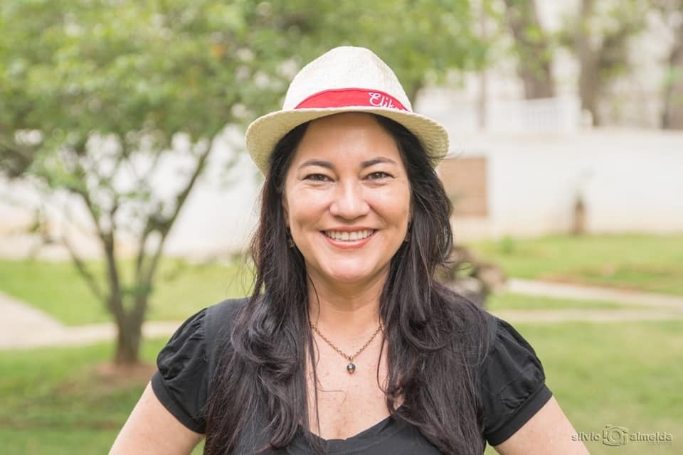 Elika Takimoto de chapéu panamá e camiseta preta, sorrindo para a câmera, com um cenário natural ao fundo