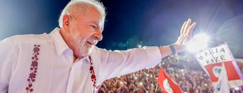 Lula veste camisa branca com adereços coloridos e acena para uma multidão reunida em evento da campanha de 2022