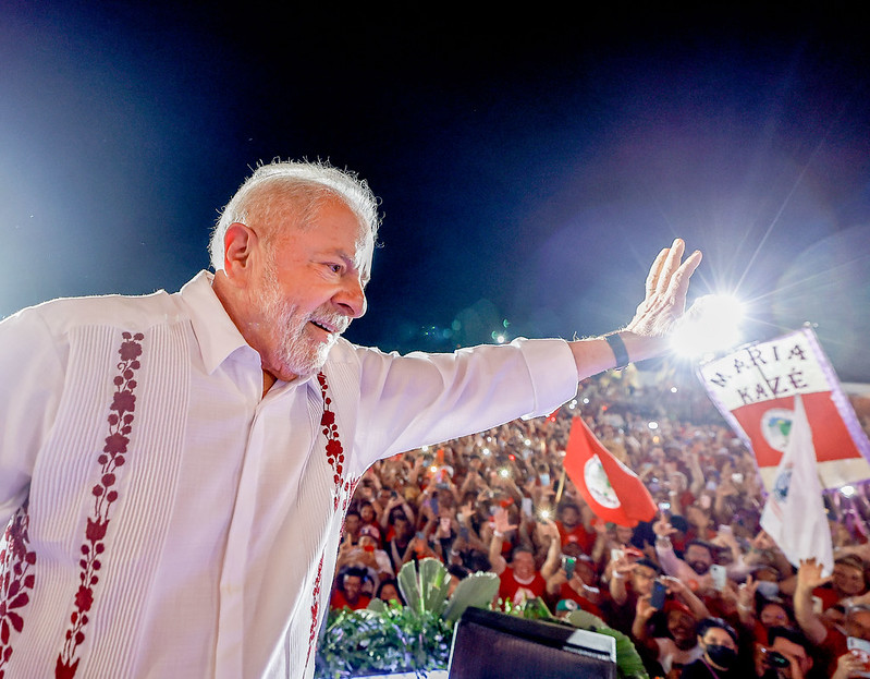Lula veste camisa branca com adereços coloridos e acena para uma multidão reunida em evento da campanha de 2022