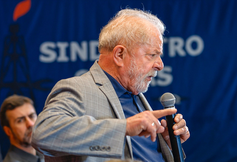 Lula segura um microfone em foto de perfil. Ele usa um terno cinza e camiseta azul marinho