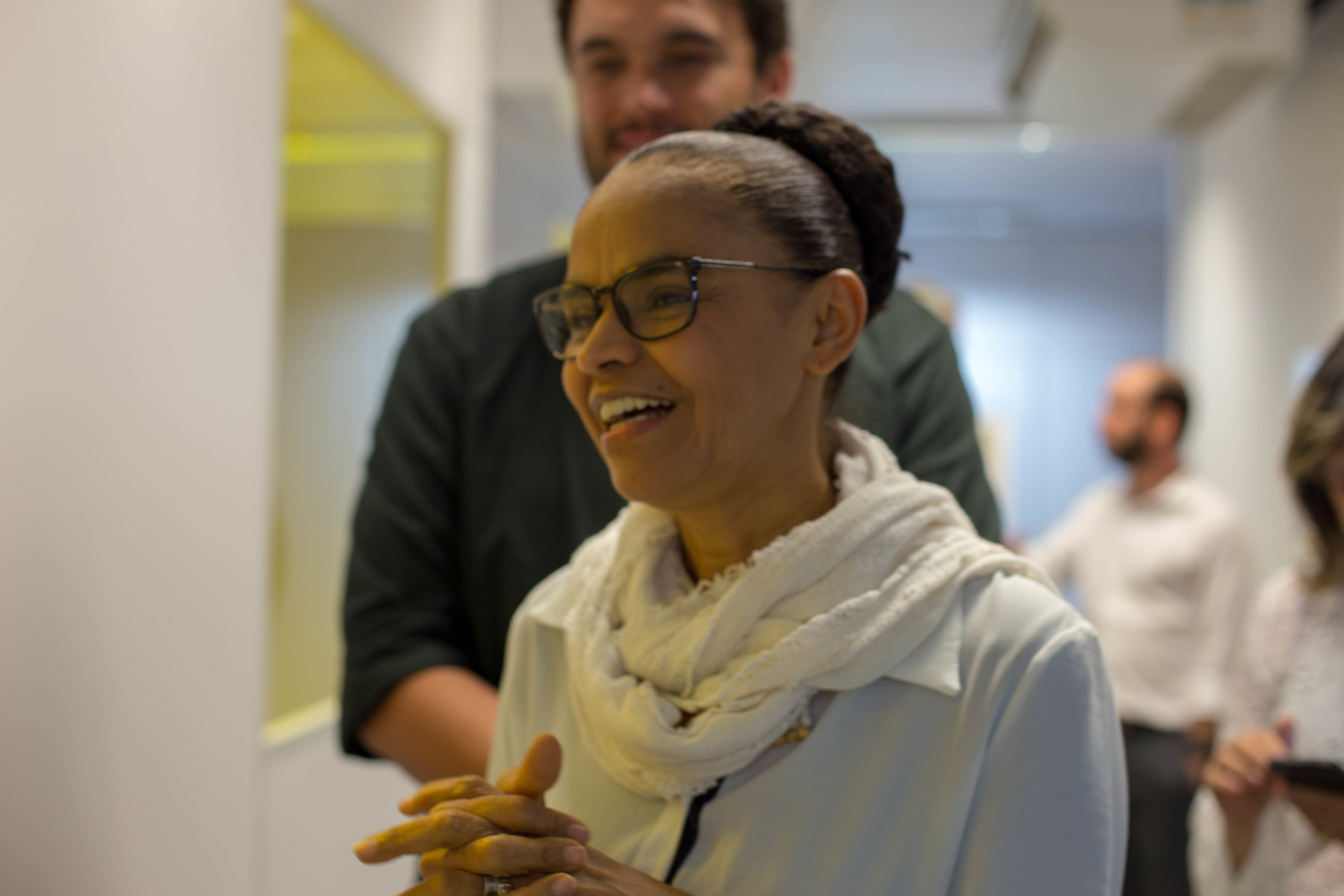 Marina Silva vesta uma blusa branca e um lenço off white no pescoço. está de óculos, sorrindo, com o cabelo preso em coque