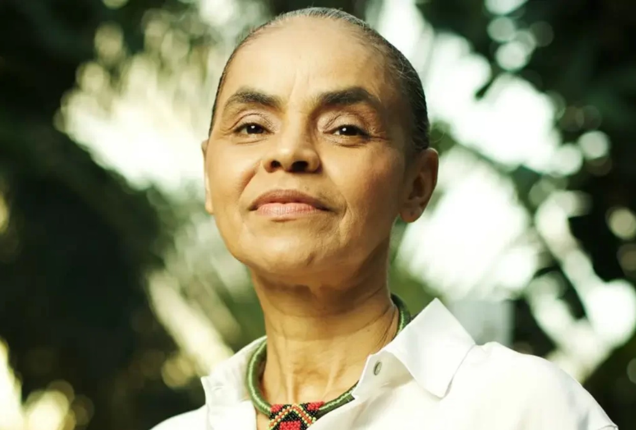 A política Marina Silva com uma camisa branca, um colar que lembra o trabalho artesanal dos povos indígenas, em foto com a natureza ao fundo