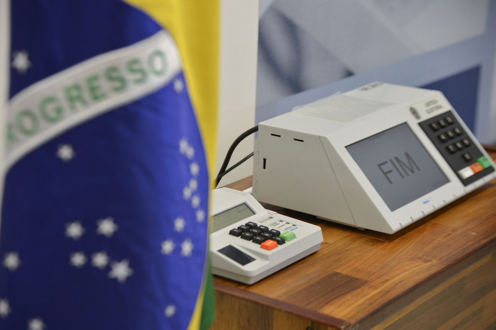 Urna eletrônica em cima de mesa marrom, ao lado da bandeira do Brasil