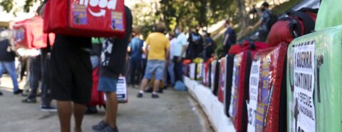 Trabalhadores do IFood enfileirados com mochilas que fazem entregas nas costas