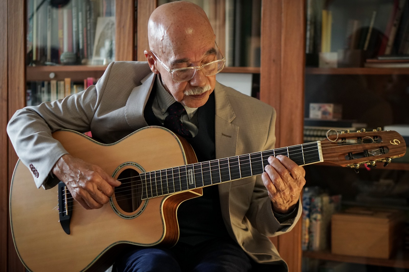 Mutinho segura e toca um violão em uma sala com livros