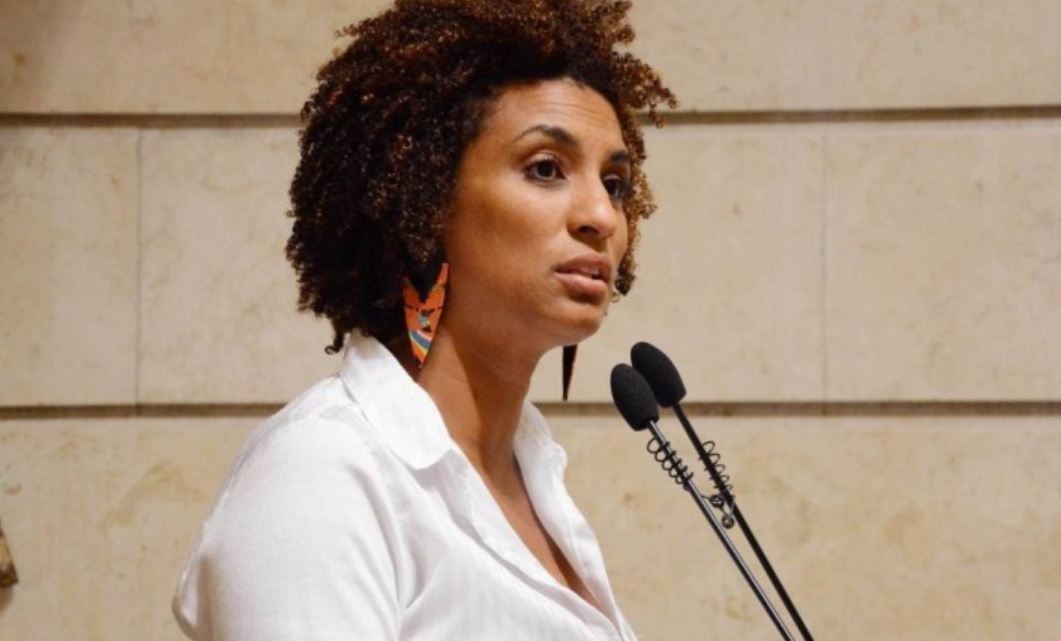 Mulher negra de perfil. Marielle Franco usa camisa branca e brincos longos, enquanto fala na tribuna da Câmara do Rio