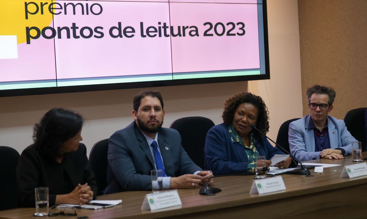 Ministra da Cultura, Margareth Menezes, conversa com outras pessoas sentada diante de uma mesa