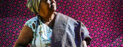 Mãe Bernadete posa para foto sentada em uma cadeira, com turbante e roupas coloridas.