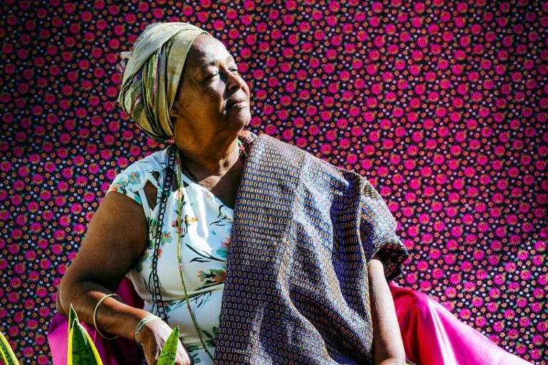 Mãe Bernadete posa para foto sentada em uma cadeira, com turbante e roupas coloridas.