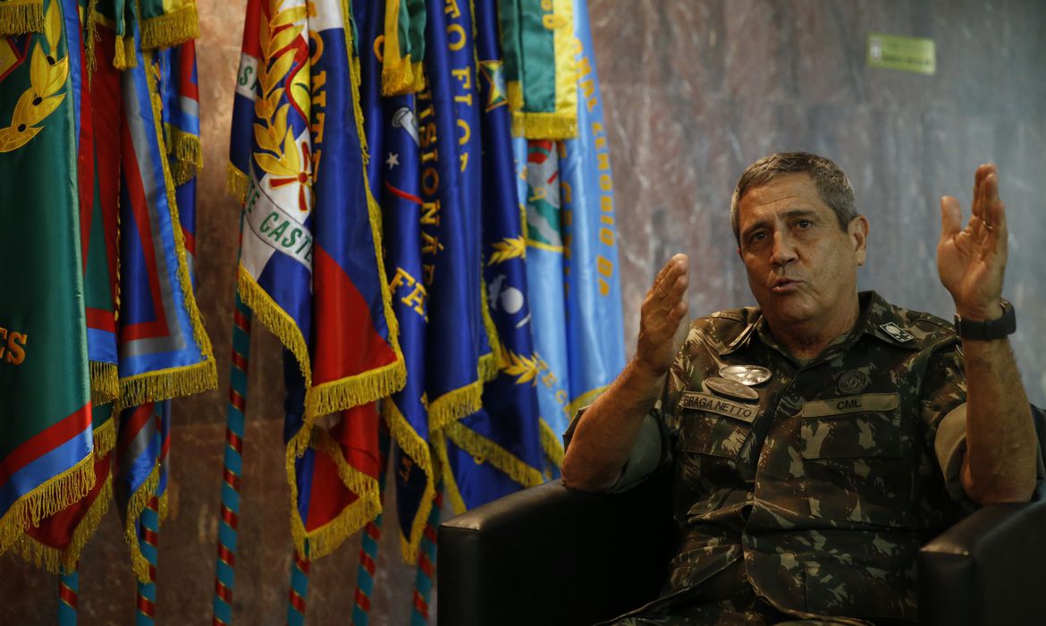 O general Walter Braga Netto é fotografado de traje militar do exército, ao lado de várias bandeiras, enquanto conversa e gesticula