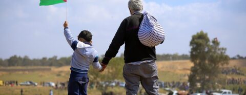 Menino e idoso palestinos olhando para o horizonte, onde há um campo com barracas ou alojamentos